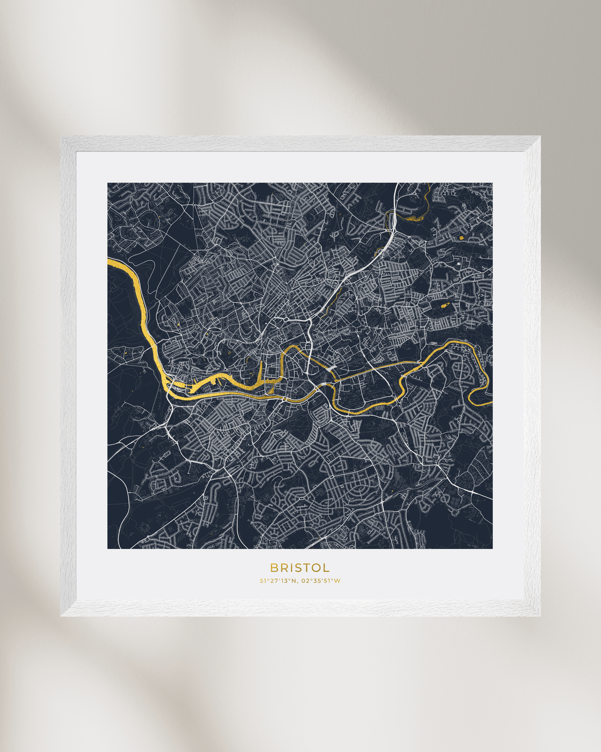 A framed minimalist city map print of Bristol, England, featuring a dark navy background with white street lines and a highlighted gold river route. The print is housed in a clean white frame and displayed against a neutral light gray background with soft, artistic shadows.
