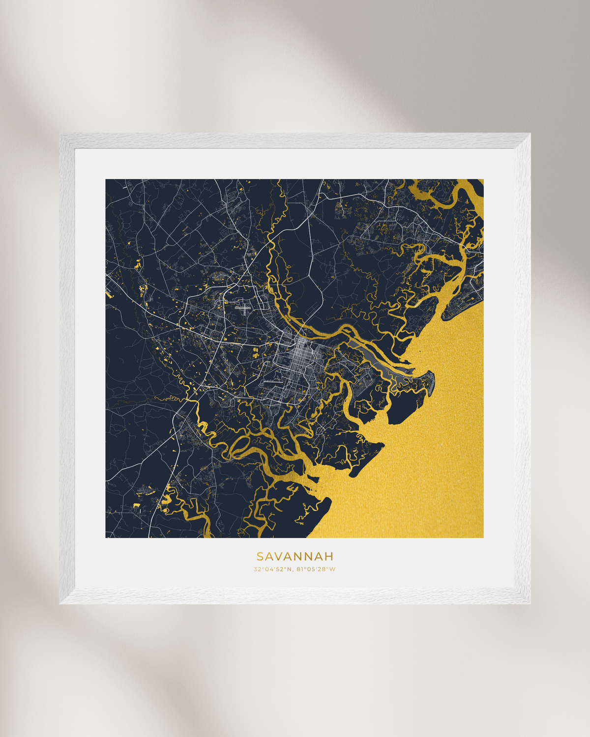 A framed minimalist map print of Savannah, Georgia, featuring a dark navy blue landmass with white road details and a textured gold coastline and water, set in a white frame against a light beige wall with soft shadows.