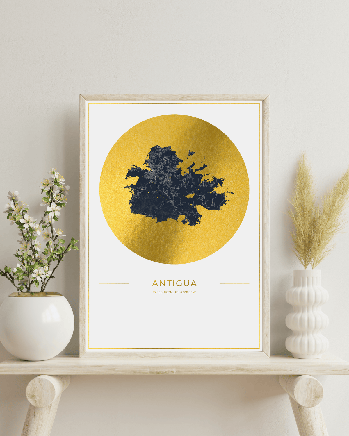 A framed art print featuring a gold foil map of Antigua, styled on a light wood shelf with white ceramic vases and dried grass.