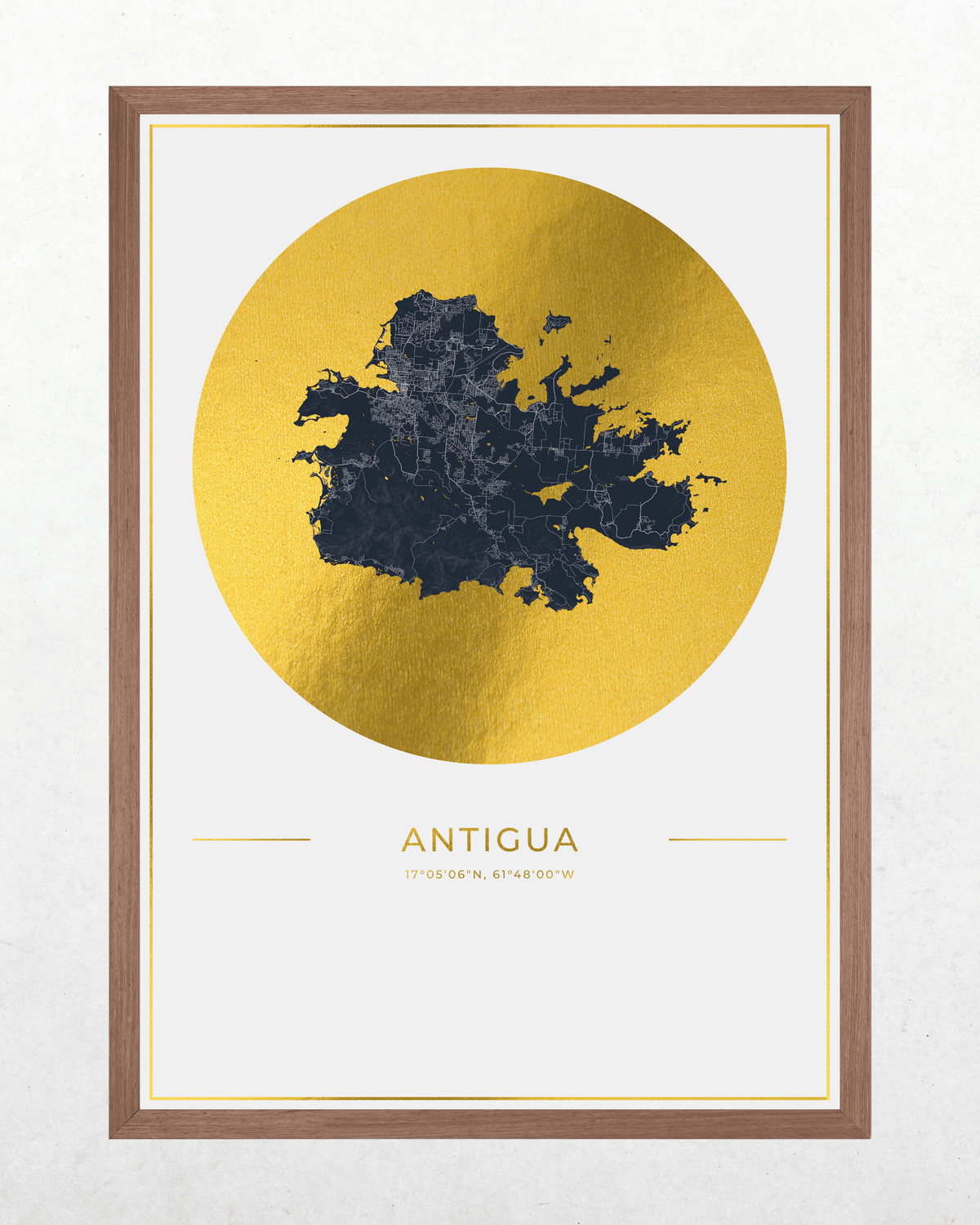 Framed wall art print featuring a dark navy map of Antigua over a large textured gold circle, with the word 'ANTIGUA' and coordinates printed below, shown in a brown wood frame against a light textured wall.