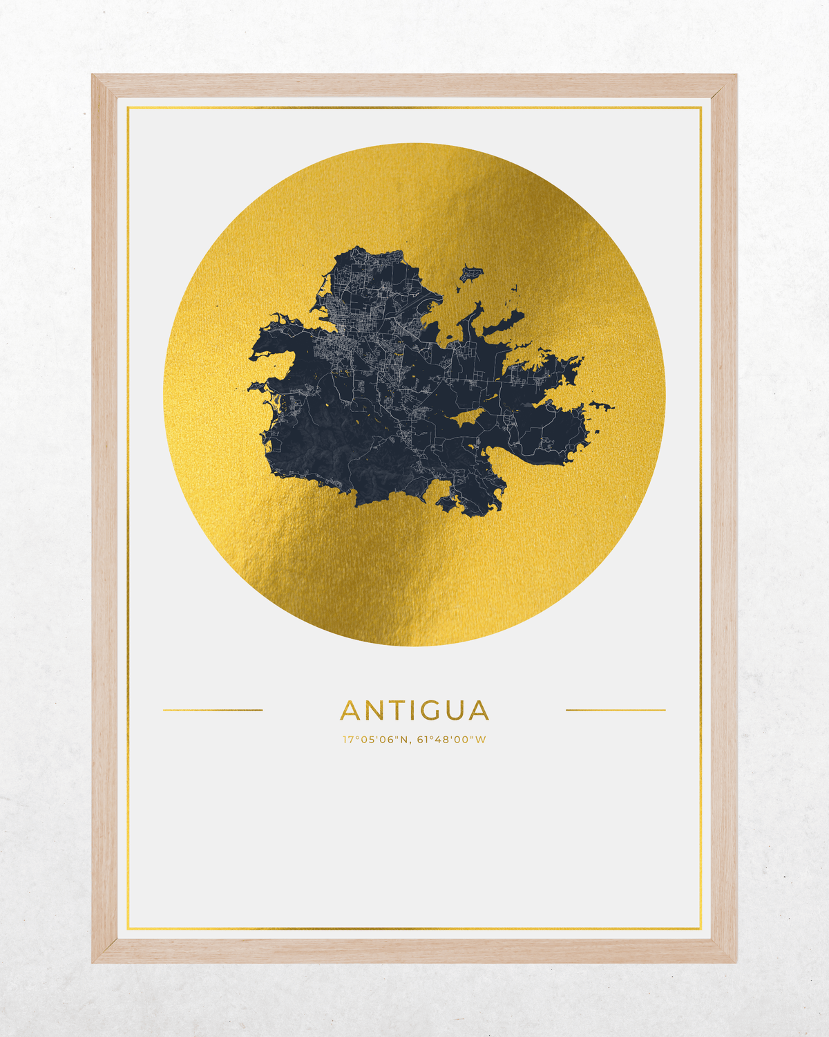 Framed wall art print featuring a dark navy map of Antigua over a large metallic gold circle, with the word 'ANTIGUA' and smaller text below, shown in a light wood frame against a white wall.
