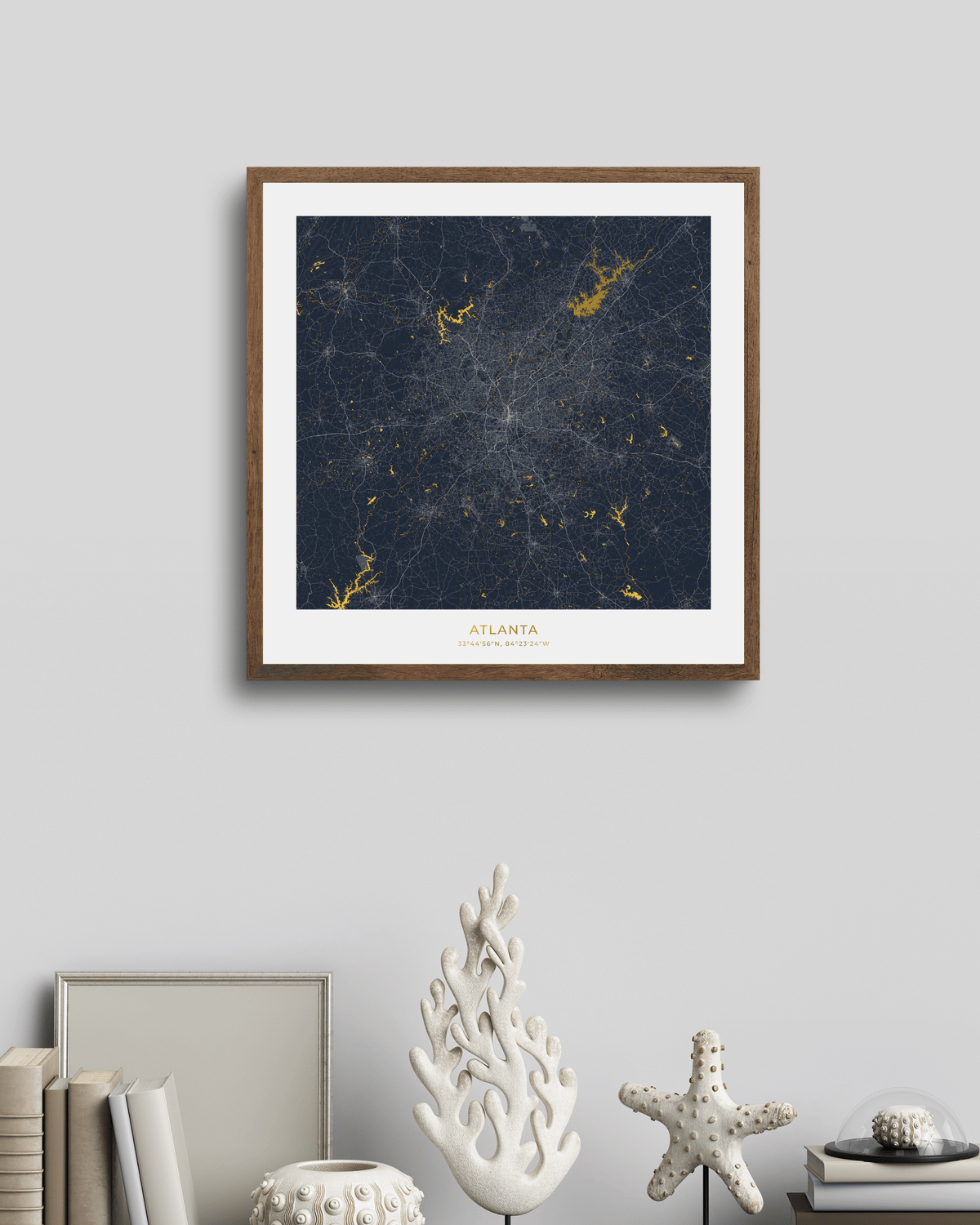 A framed city map print of Atlanta, Georgia, featuring a dark navy blue background with gold waterways & lakes and a white mat border, displayed on a light gray wall above a styled shelf with books and decorative objects.