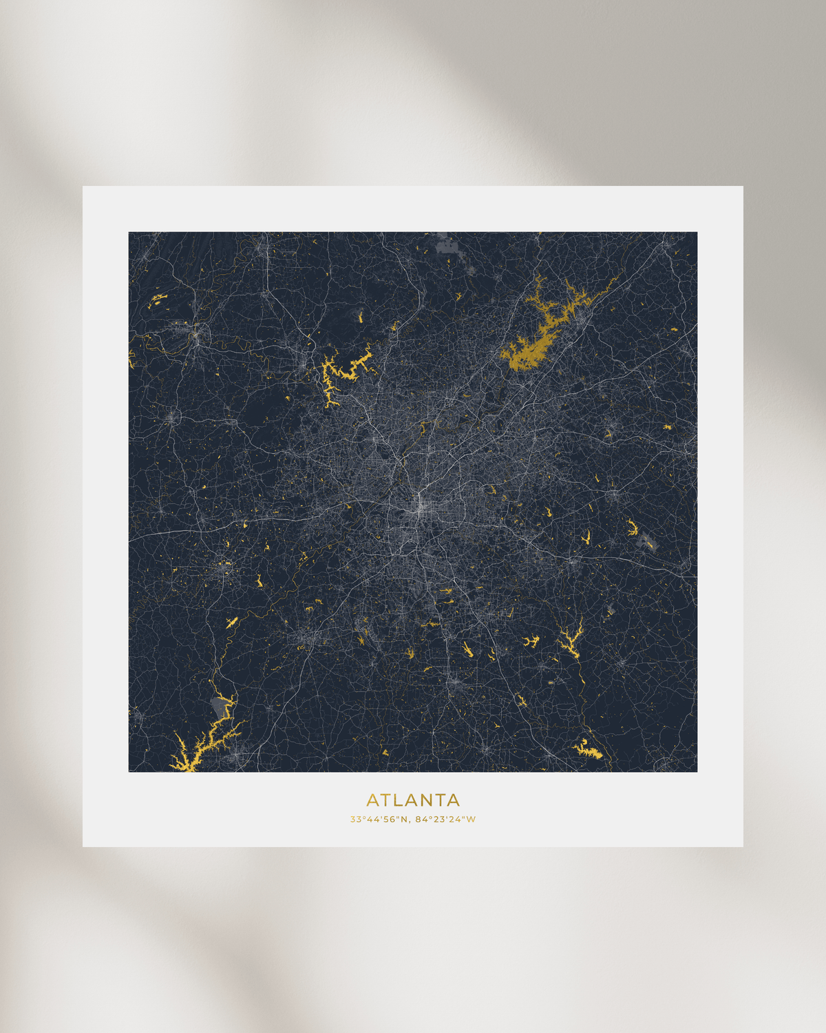 A framed city map print of Atlanta, featuring a dark navy background with gold waterways and lakes and white road lines, centered on a light beige wall with soft shadows.