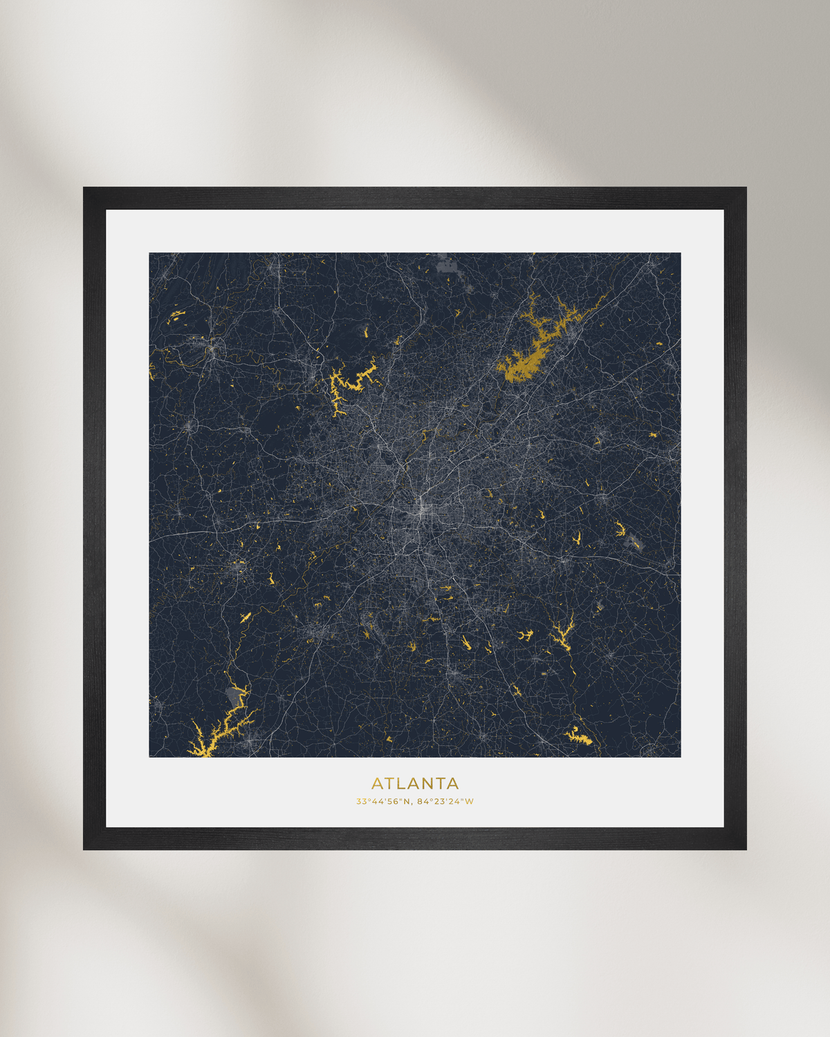 A framed city map print of Atlanta, Georgia, featuring a dark navy blue background with gold waterways & lakes and white street lines, surrounded by a white mat and a simple black frame against a light gray wall.