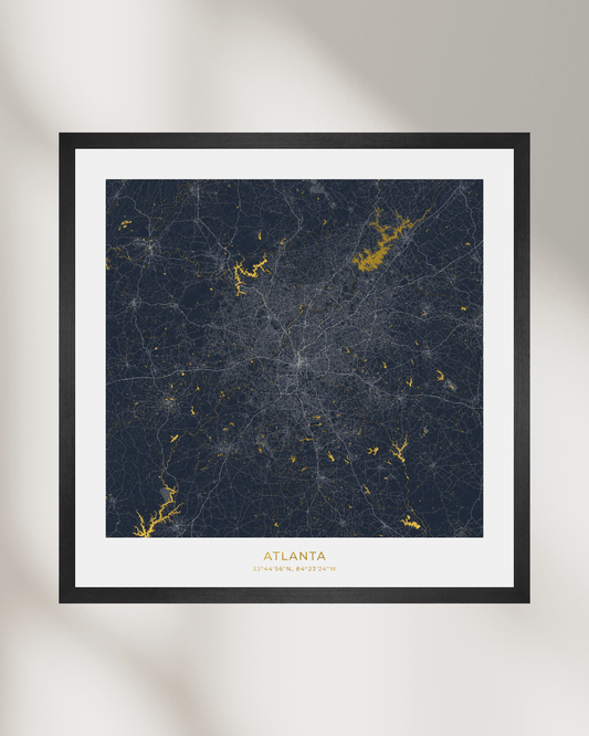 A framed city map print of Atlanta, Georgia, featuring a dark navy blue background with gold waterways & lakes and white street lines, surrounded by a white mat and a simple black frame against a light gray wall.