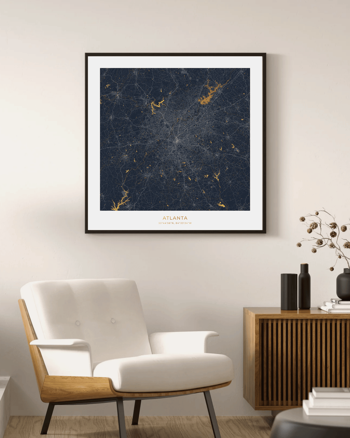 A framed art print of the Atlanta city map with a dark navy blue background and gold waterways & lakes , displayed on a beige wall above a modern white armchair and wooden sideboard.
