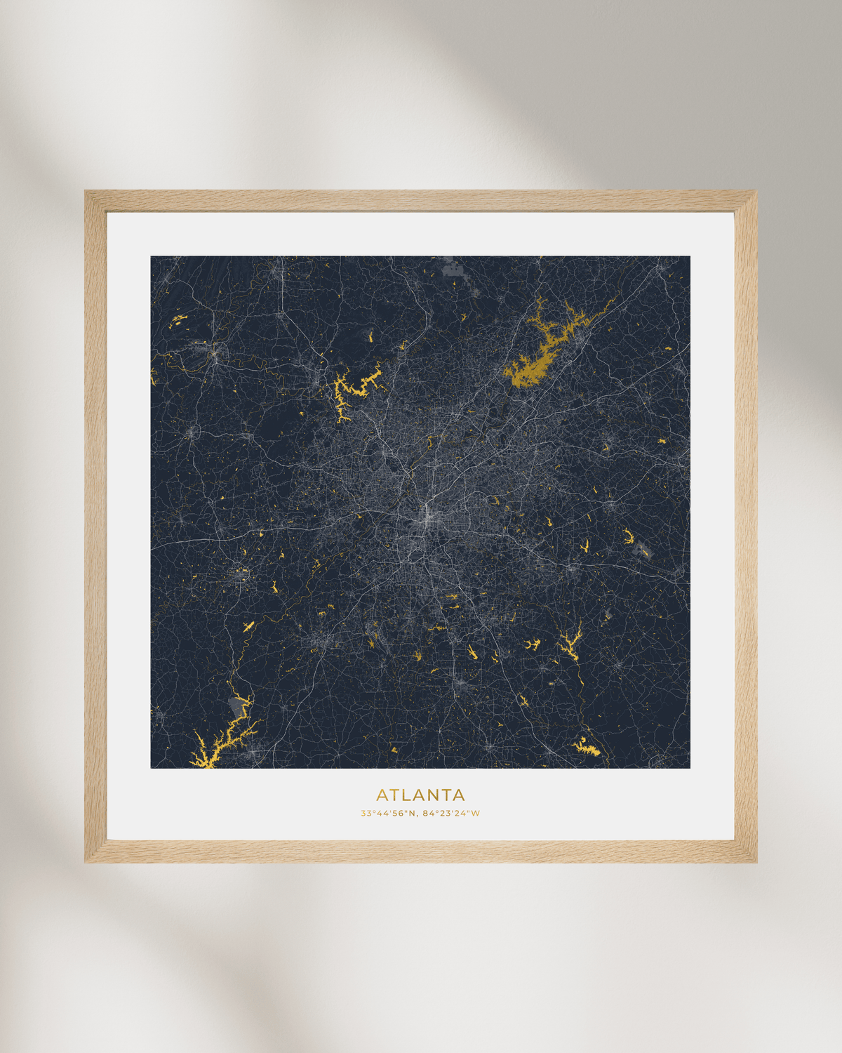 A framed city map print of Atlanta, Georgia, featuring a dark navy blue background with gold waterways & lakes, surrounded by a white mat and a light natural wood frame, displayed against a neutral light gray wall with soft shadows.
