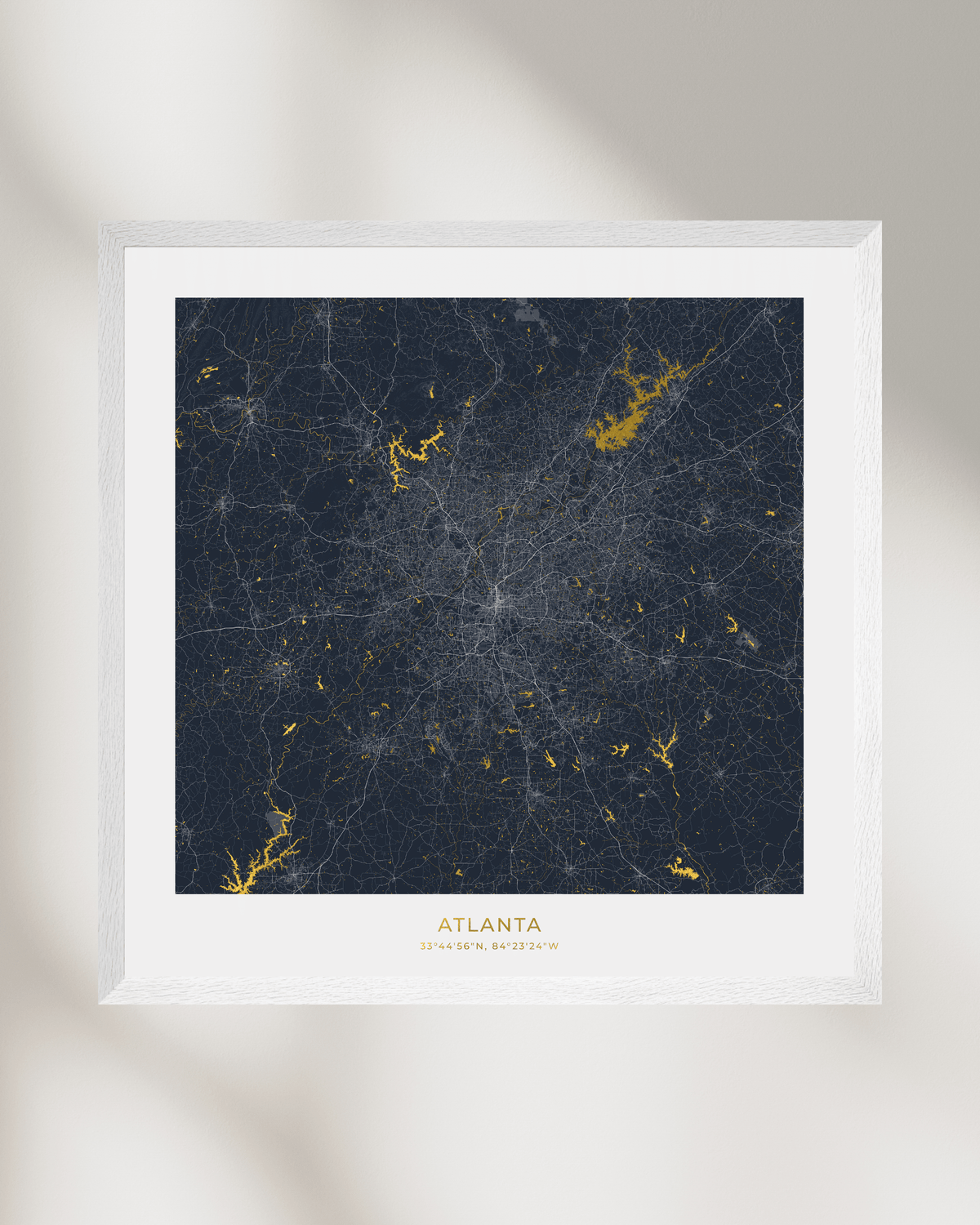A framed city map print of Atlanta, Georgia, featuring a dark navy blue background with gold waterways & lakes, surrounded by a white mat and a white wood frame, displayed against a neutral light gray wall with soft shadows.