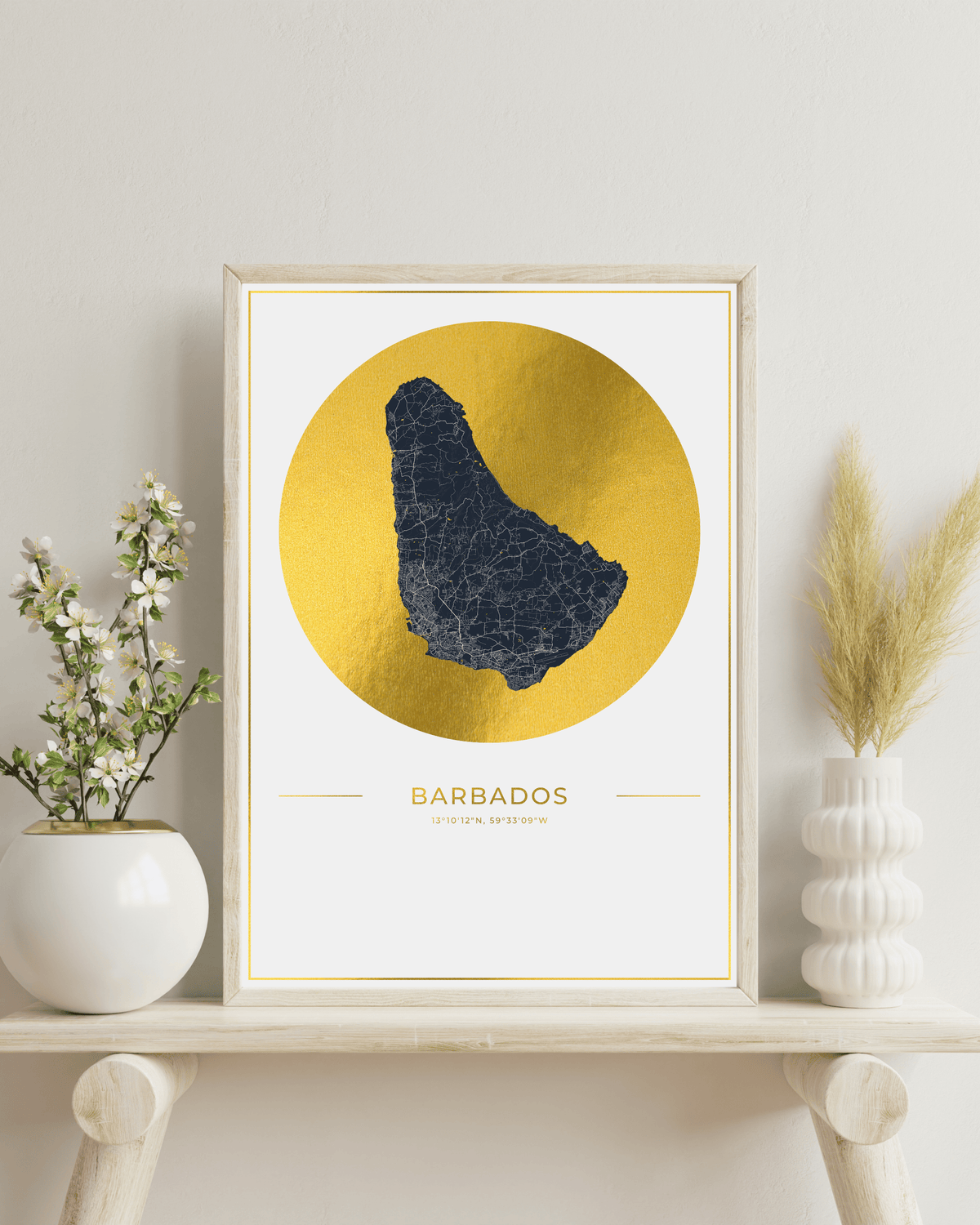 A framed art print featuring a dark blue map of Barbados over a large gold circle on a white background, styled on a light wood shelf with decorative vases and dried grass.
