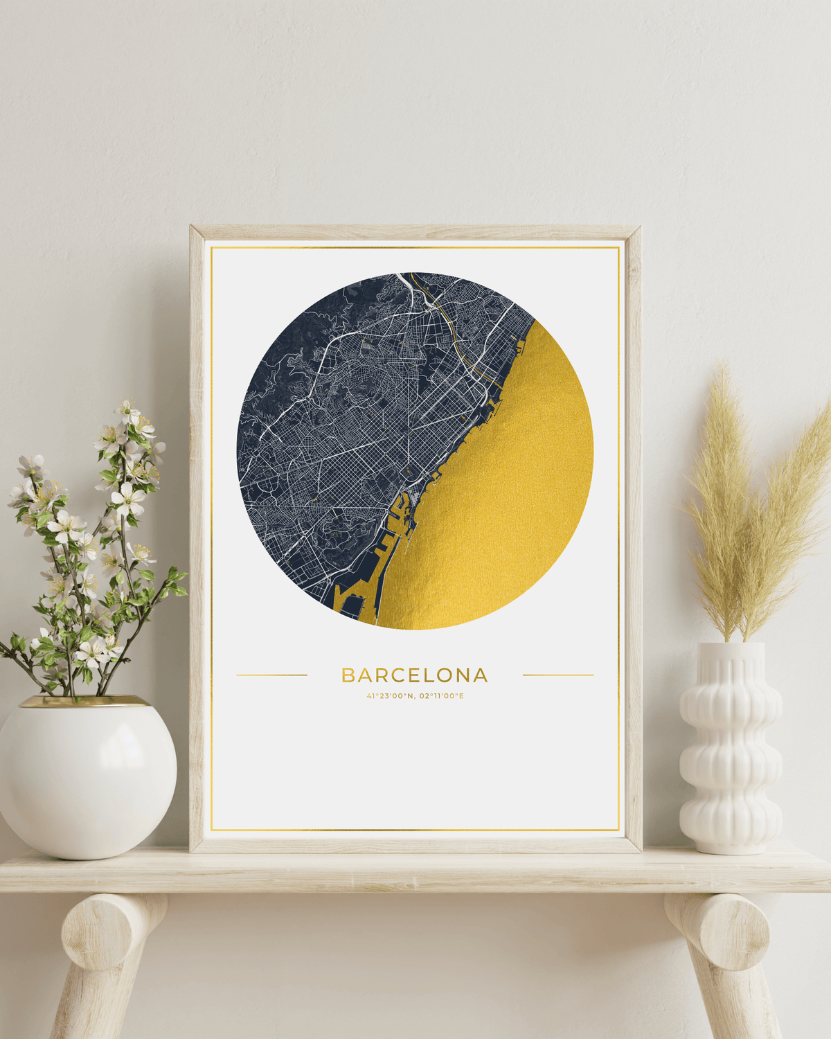 A framed minimalist Barcelona city map art print with a navy and gold circular design, displayed on a light wood shelf with decorative white vases and dried grass.