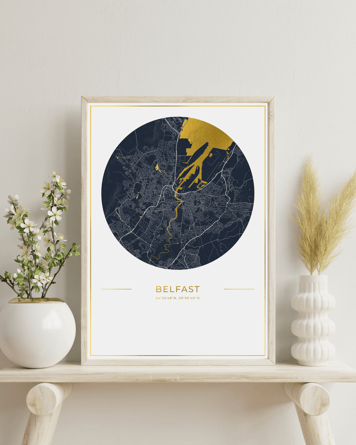 A framed city map print of Belfast, Ireland, featuring a circular dark navy map with gold river and coastline highlights, a white mat, and a black frame on a light textured background.