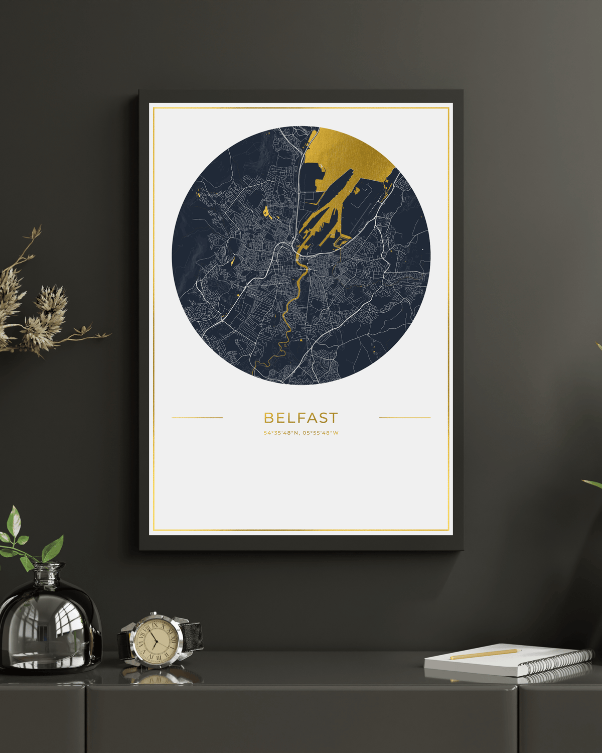 A framed circular Belfast city map print hangs on a dark charcoal wall above a console table. The map features a navy blue and gray city layout with a prominent gold coastal area and gold text reading 'BELFAST' below.