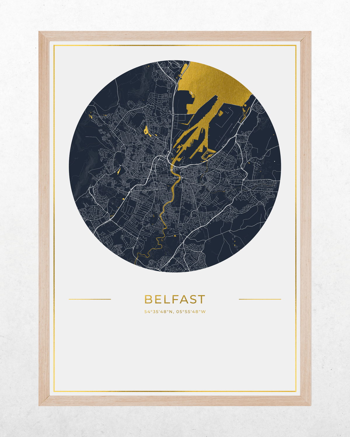 A framed minimalist city map print of Belfast, featuring a circular navy and gold map design with the name'BELFAST' printed below, set in a light wood frame against a textured off-white wall.