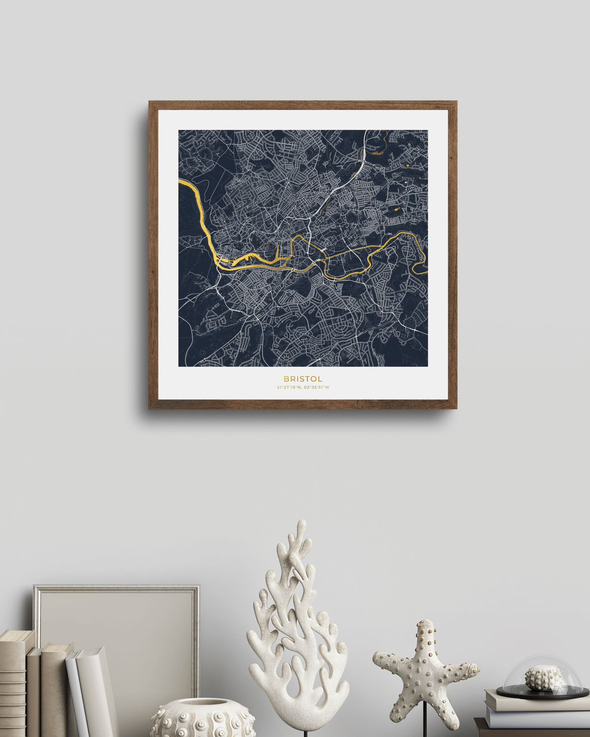 A framed city map print of Bristol, England, featuring a dark navy background with light gray street details and a bright gold river line. The print is housed in a natural wood frame with a white mat, hanging on a light gray wall above a styled shelf with books and decorative objects.