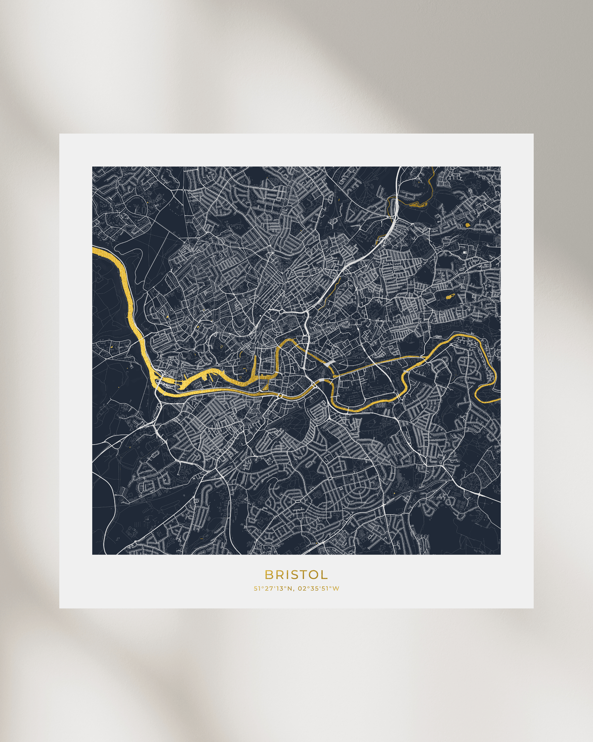 A framed-style art print featuring a dark navy street map of Bristol with a bright gold highlighted river route and white grid lines. The print has a wide white border and the word BRISTOL printed at the bottom, shown against a softly lit neutral background.