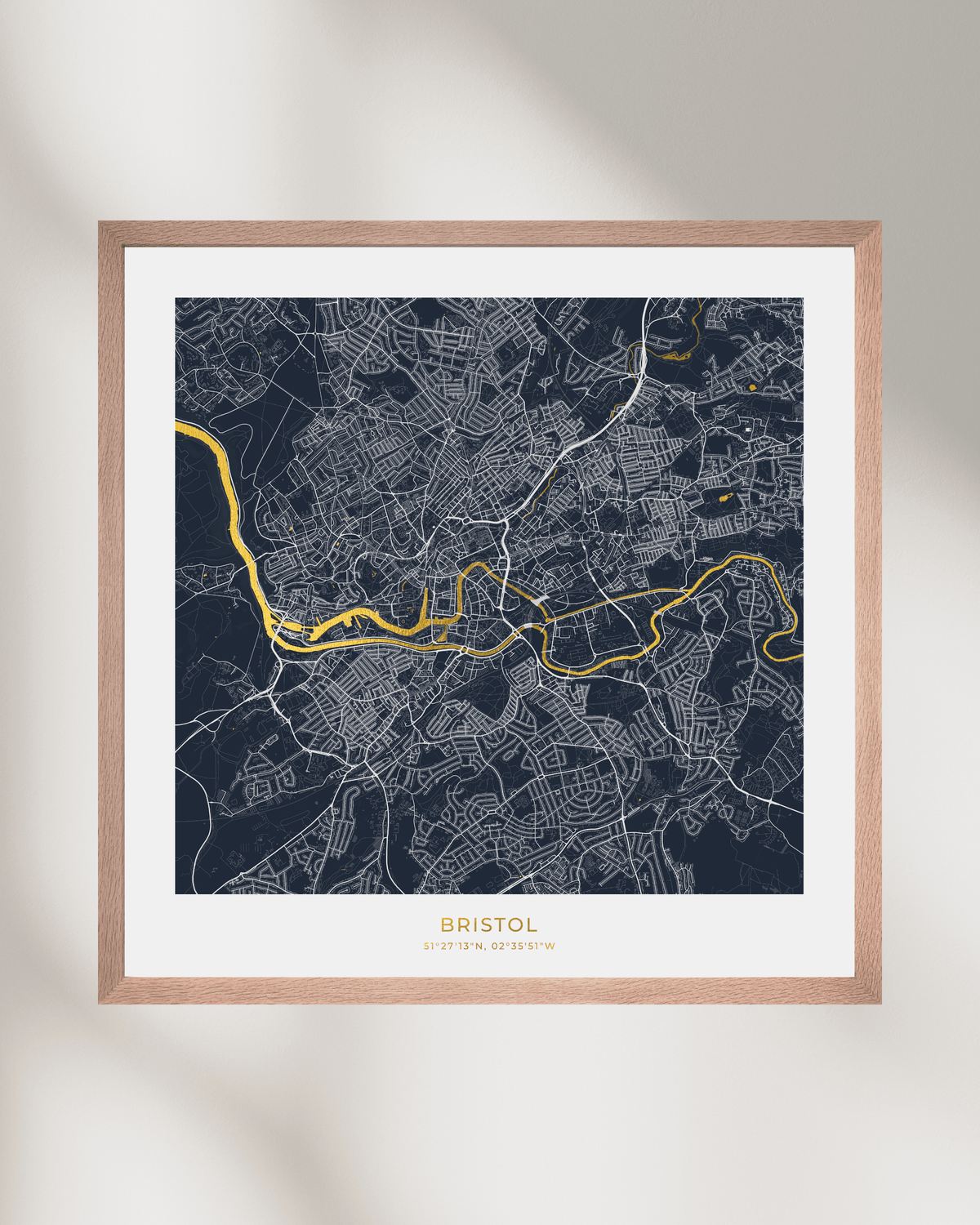 A framed city map print of Bristol, England, featuring a dark navy background with white street lines and a bright gold highlighted river route. The print is matted in white and enclosed in a light natural wood frame, shown hanging on a light beige wall with soft shadows.