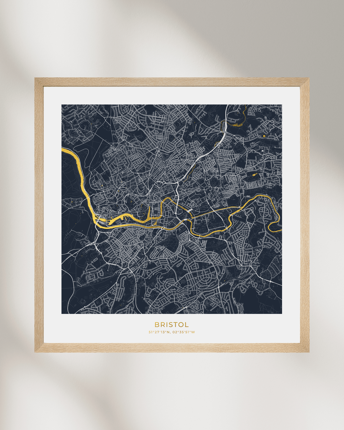 A framed city map print of Bristol, England, featuring a dark navy background with white street lines and a highlighted gold River Avon. The print is matted in white and enclosed in a light natural wood frame, shown hanging on a light gray wall with soft shadows.