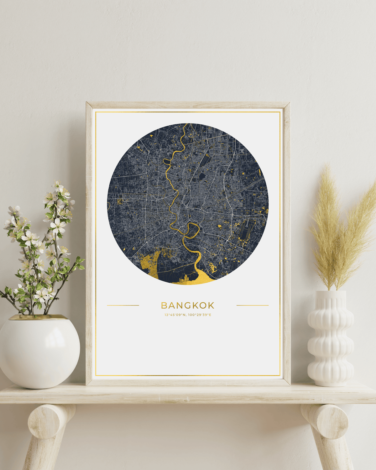 A framed circular map print of Bangkok, Thailand, featuring a dark gray street layout with gold highlights and the city name printed below, shown in a light wood frame against a white wall.