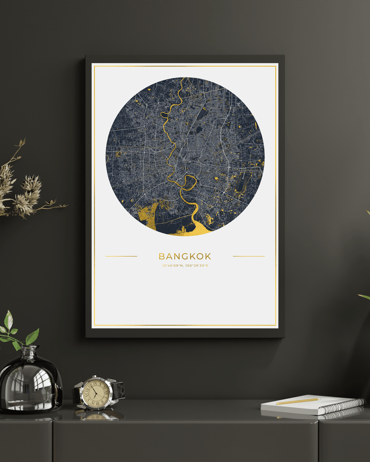 A framed circular map print of Bangkok, Thailand, featuring a dark navy blue street pattern with gold highlights on an off-white background. The print is housed in a black frame and is displayed on a dark gray wall above a console table decorated with a glass bottle, a clock, and a notebook.