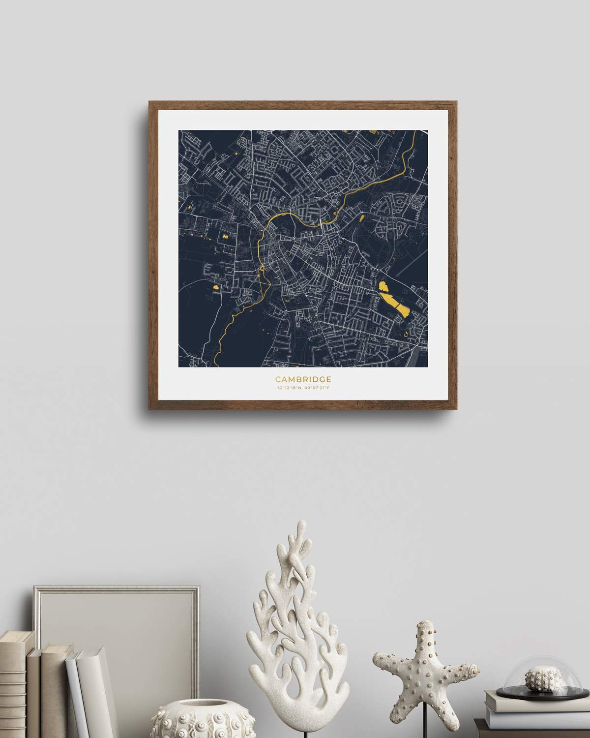 A framed square map print of Cambridge, UK, featuring a dark navy blue background with white street lines and gold highlights, displayed on a light gray wall above a styled shelf with books and decorative objects.