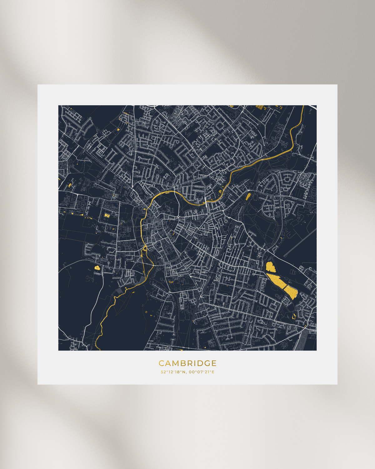 A minimalist city map print of Cambridge, UK, featuring a dark navy blue background with light gray street lines and bright gold highlights for roads and landmarks. The print is presented on a white cardstock with the city name and coordinates printed at the bottom, set against a warm beige background with soft shadows.