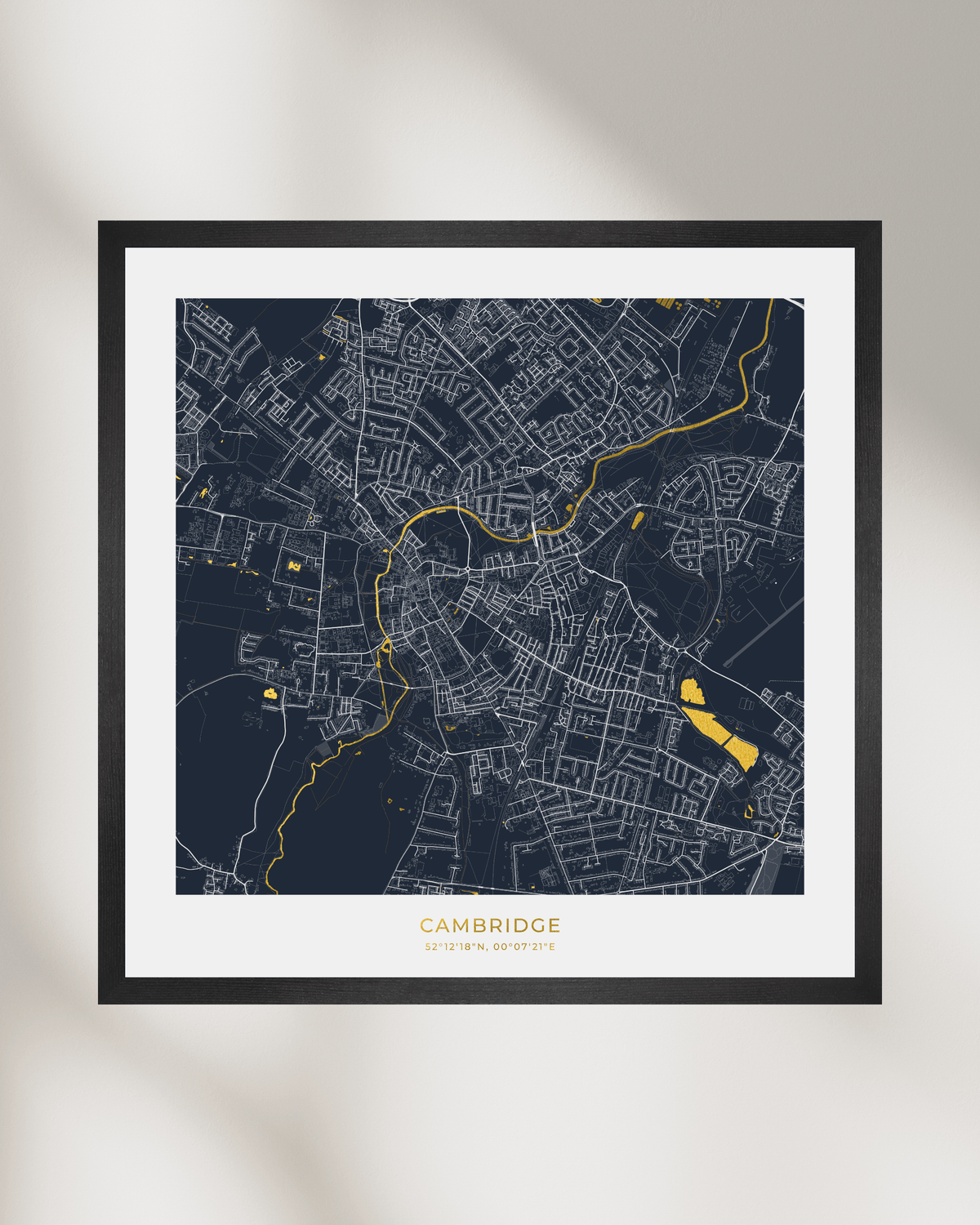 A framed city map print of Cambridge, UK, featuring a dark navy background with white street lines and bright gold highlights, displayed on a light beige wall with soft shadows.