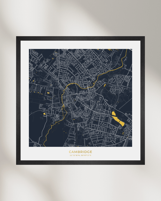 A framed city map print of Cambridge, UK, featuring a dark navy background with white street lines and bright gold highlights, displayed on a light beige wall with soft shadows.