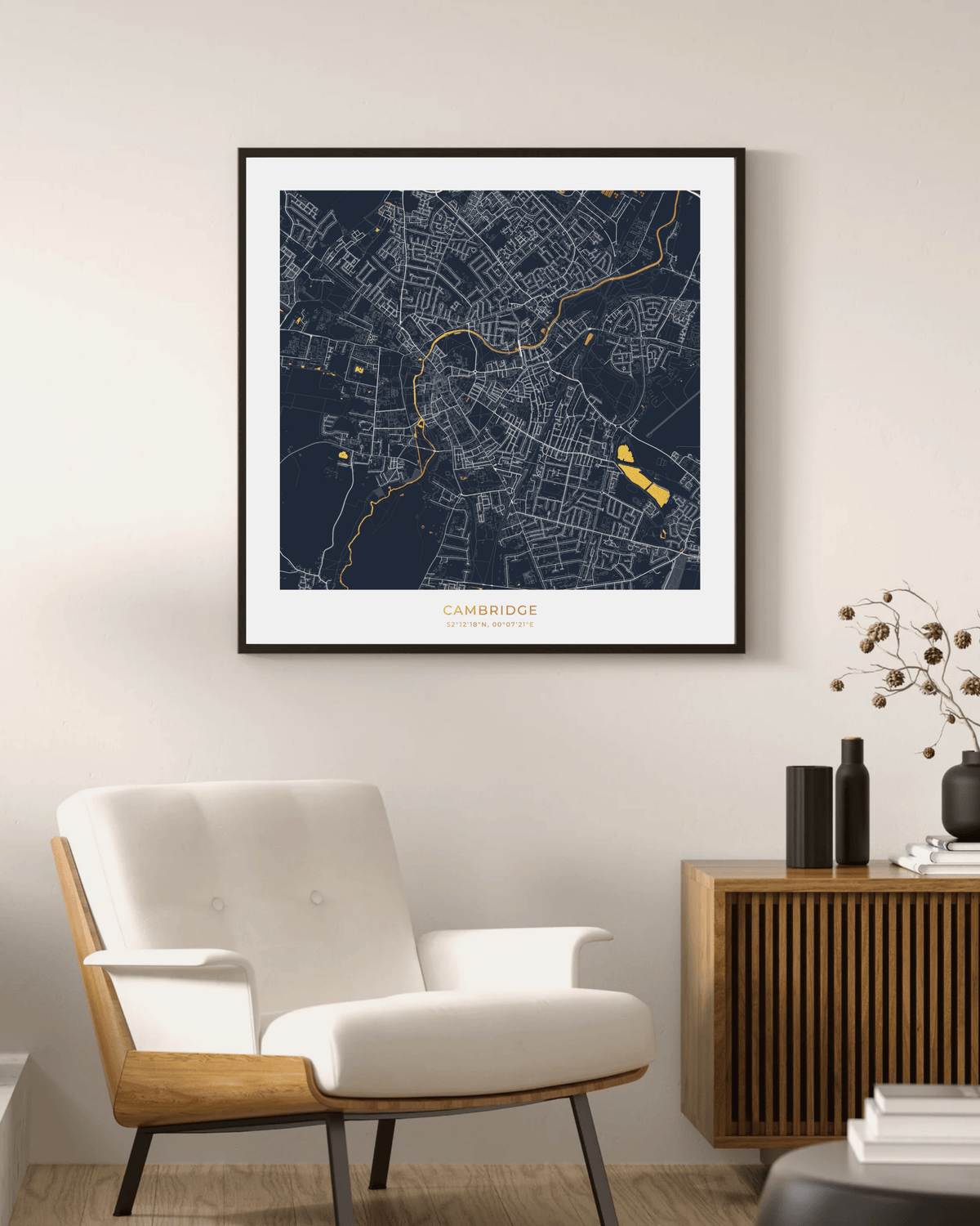 A framed art print of a Cambridge city map, featuring a dark navy blue background with white street lines and gold highlights, displayed on a beige wall above a modern white armchair and wooden sideboard.