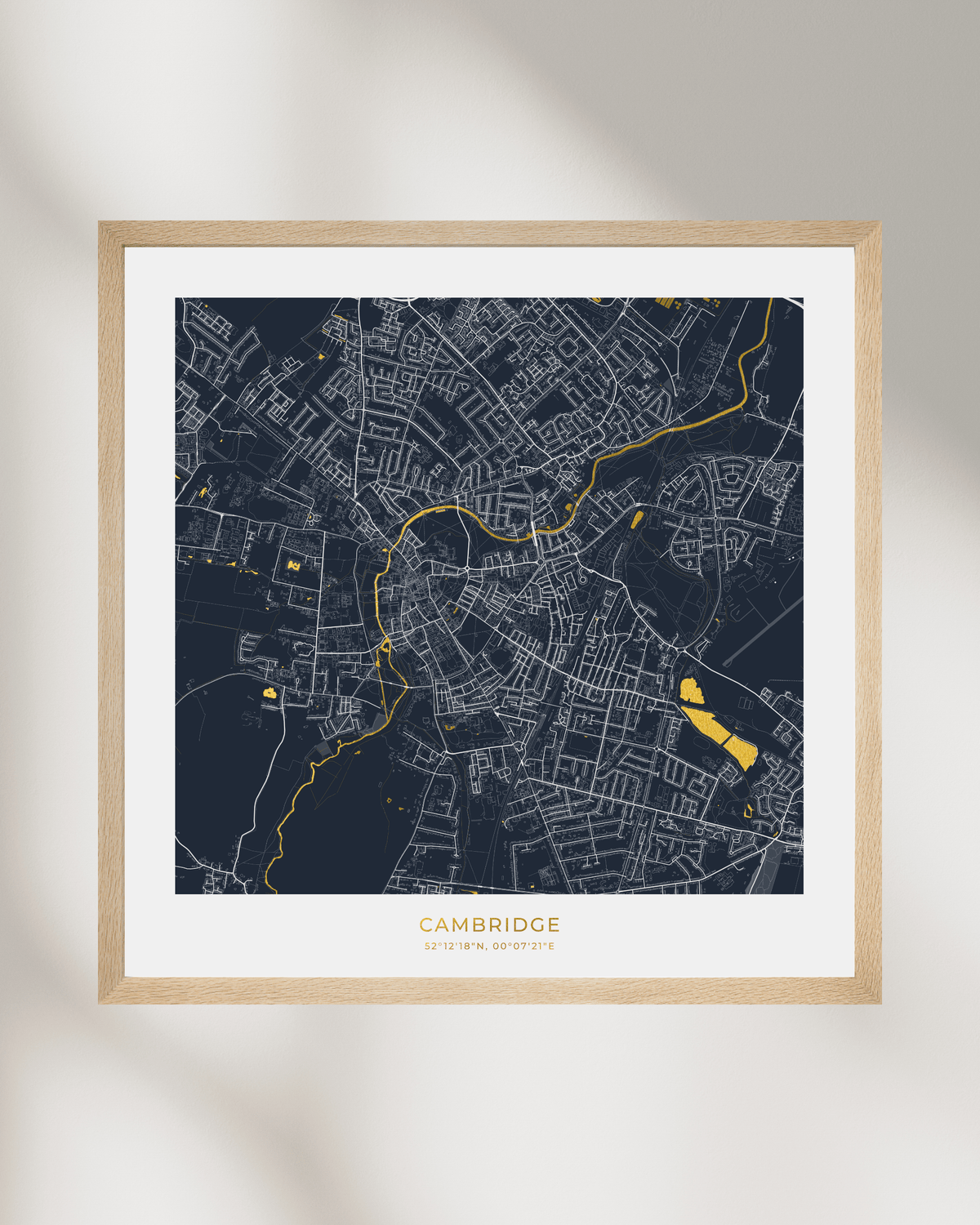 A framed city map print of Cambridge, UK, featuring a dark navy background with white street lines and bright gold highlights, displayed on a light beige wall with soft shadows.