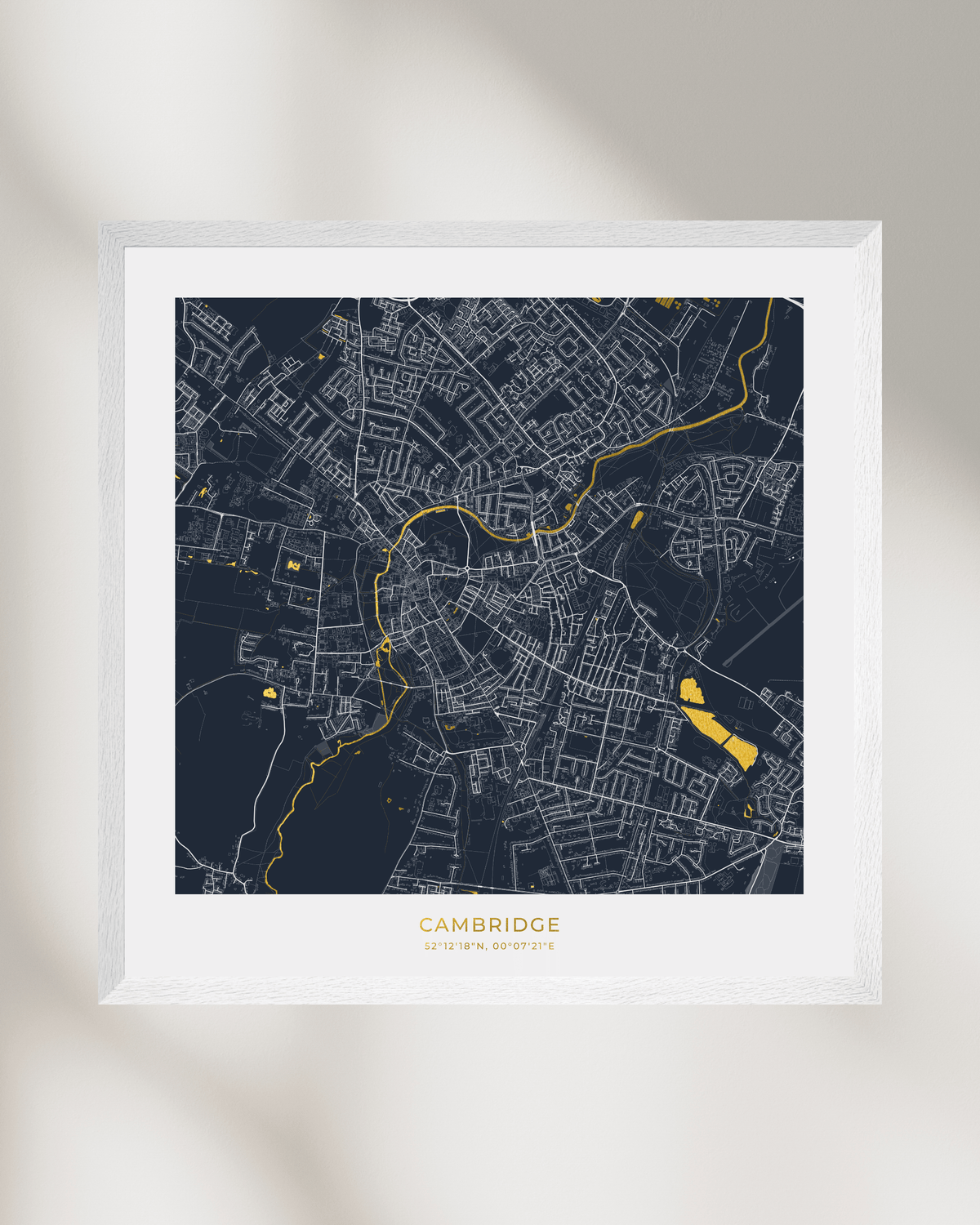 A framed city map print of Cambridge, UK, featuring a dark navy background with white street lines and bright gold highlights, displayed on a light beige wall with soft shadows.