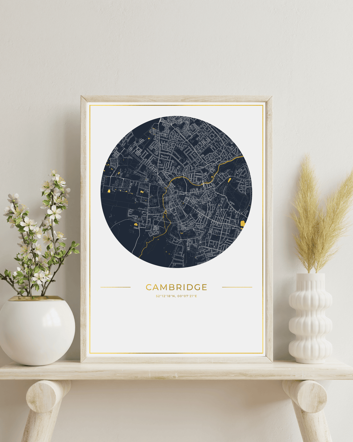 A framed circular city map print of Cambridge, UK, featuring a dark navy street network with gold line details on a white background, with the word 'CAMBRIDGE' and smaller text below, shown in a black frame against a light textured wall.