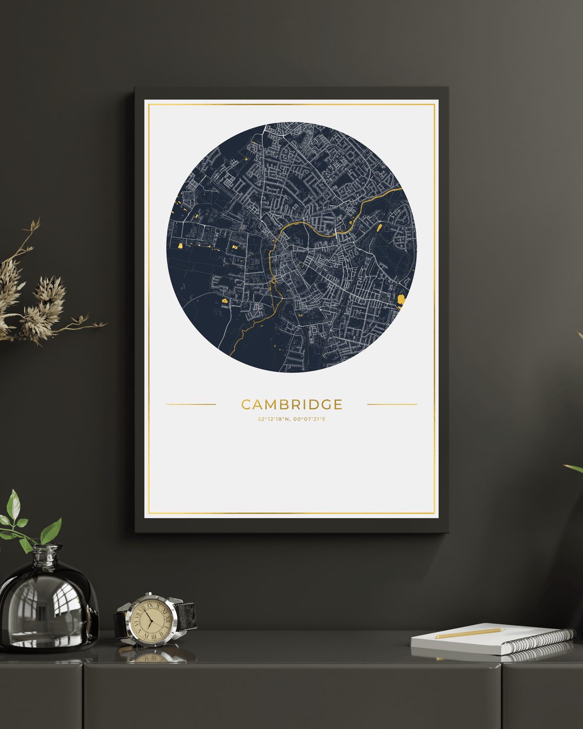A framed circular city map print of Cambridge, UK, featuring a dark navy street network with gold line details on a white background, with the word 'CAMBRIDGE' and smaller text below, shown in a black frame against a light textured wall.