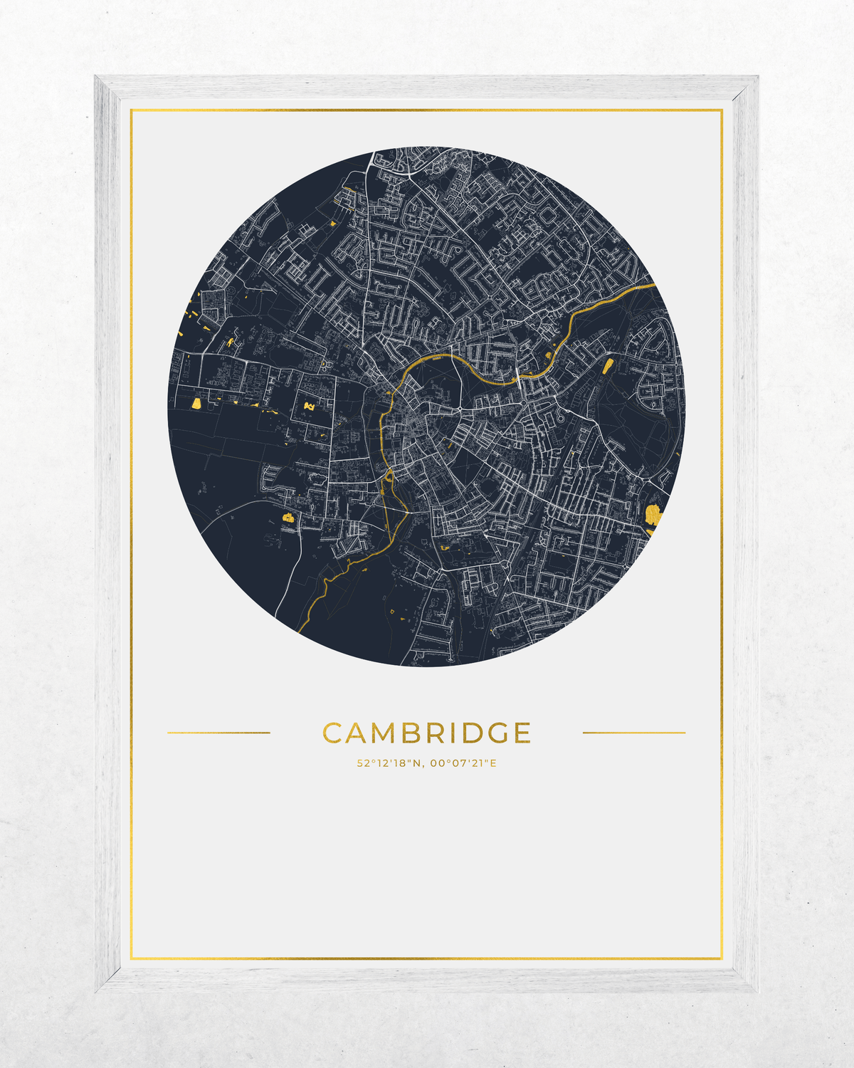 A framed circular city map print of Cambridge, featuring a dark navy map with fine white street lines and small gold highlights, centered on a white background with the word CAMBRIDGE and small coordinates below. The print is shown in a light wood frame against a textured white wall.