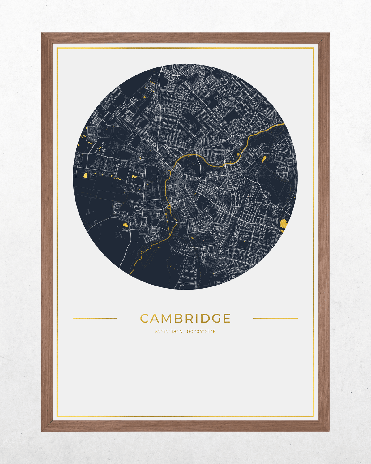 A framed circular city map print of Cambridge, UK, featuring a dark navy street network with gold line details on a white background, with the word 'CAMBRIDGE' and smaller text below, shown in a brown wood frame against a light textured wall.