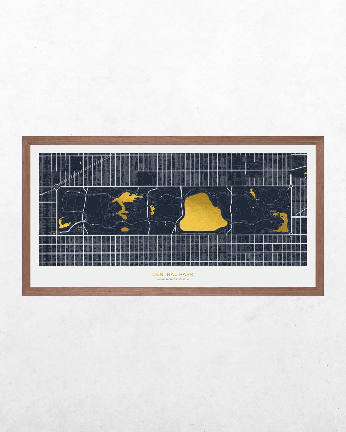 A framed art print of a stylized street map of Central Park, New York City, featuring dark navy blue land, bright gold water features, and a white mat board inside a light brown wooden frame, displayed on a textured white wall.