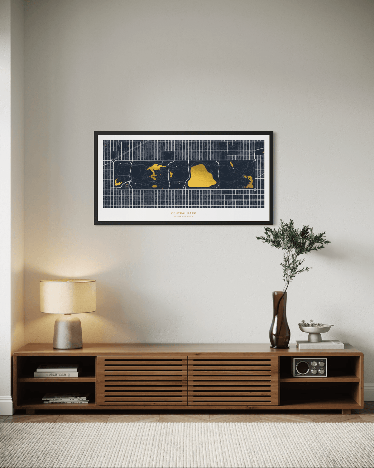 A framed horizontal city map print of Central Park with a black and navy grid and bright gold highlighted areas, displayed on a light gray wall above a wooden media console in a styled interior setting.