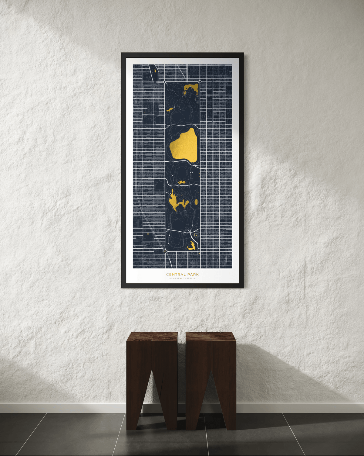 A framed vertical map print of New York City hangs on a textured white wall above two dark wooden stools. The print features a dark navy street grid with bright gold highlighted boroughs and white text at the bottom.