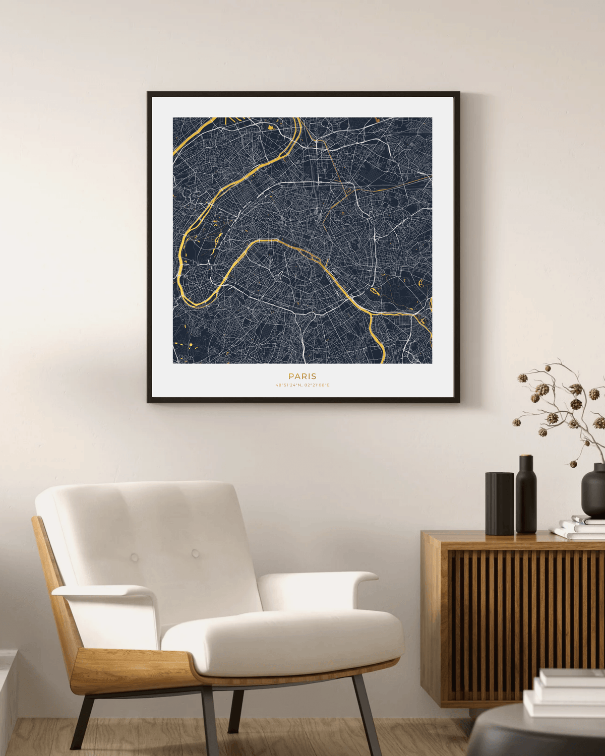 A framed Paris city map art print with a dark navy background, white street lines, and gold river routes, hanging on a beige wall above a modern white armchair and wooden sideboard.
