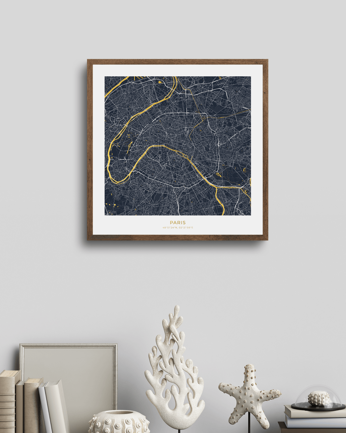 A framed Paris city map art print with a dark navy background, gold street lines, and a white mat inside a brown wooden frame, displayed on a light gray wall above a styled shelf with books and decorative objects.