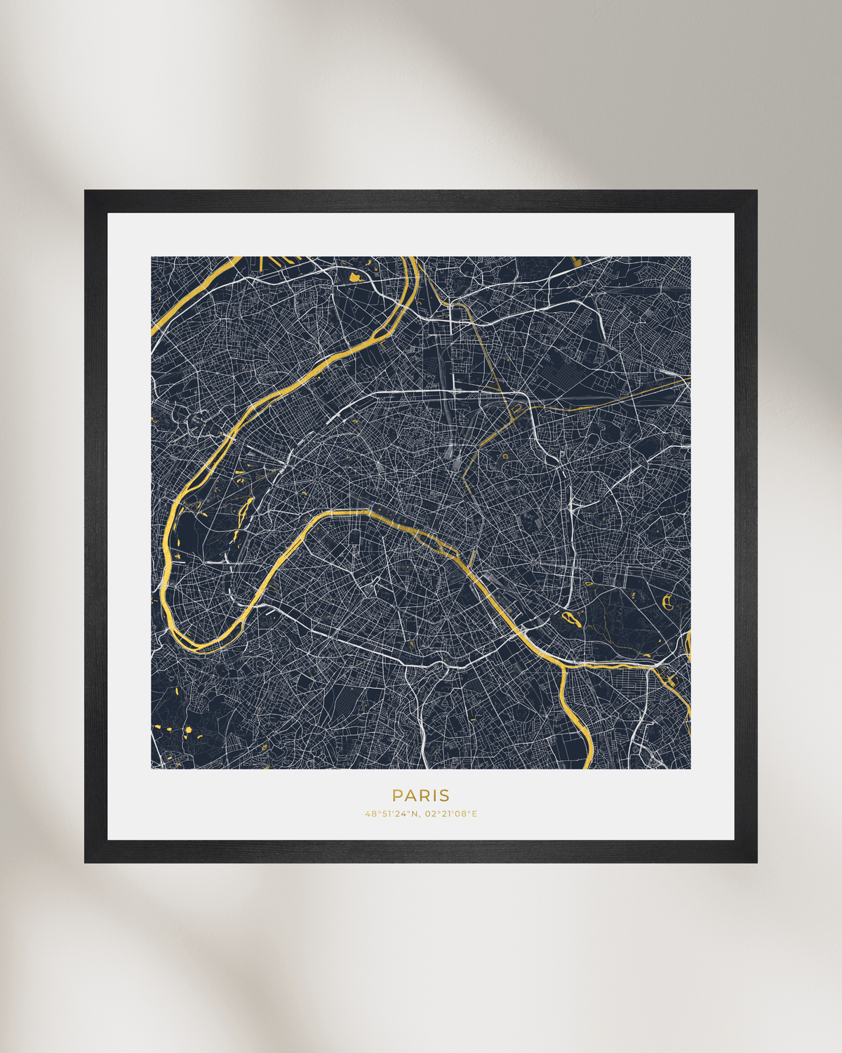 A framed Paris city map art print with a dark navy background, white street lines, and gold river highlights, displayed against a light gray wall with soft shadow overlays.