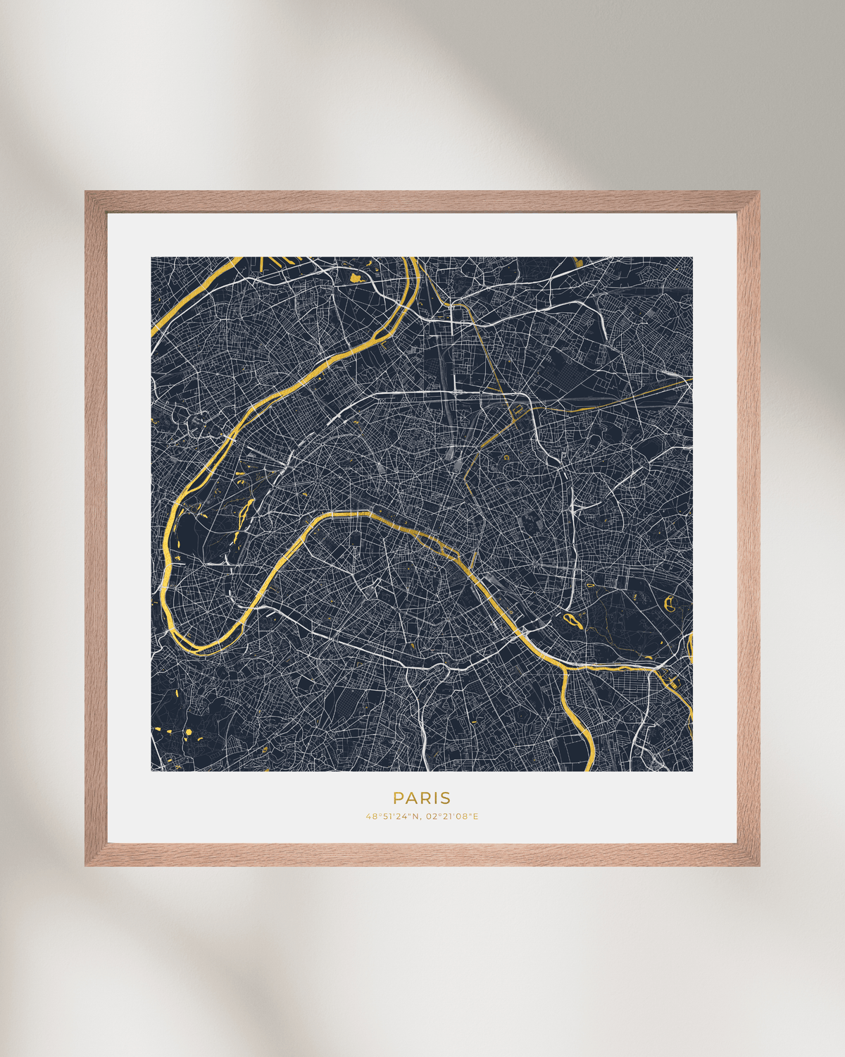 A framed Paris city map art print with a dark navy background, white street lines, and gold river and text details, set in a light wood frame against a warm off-white wall.