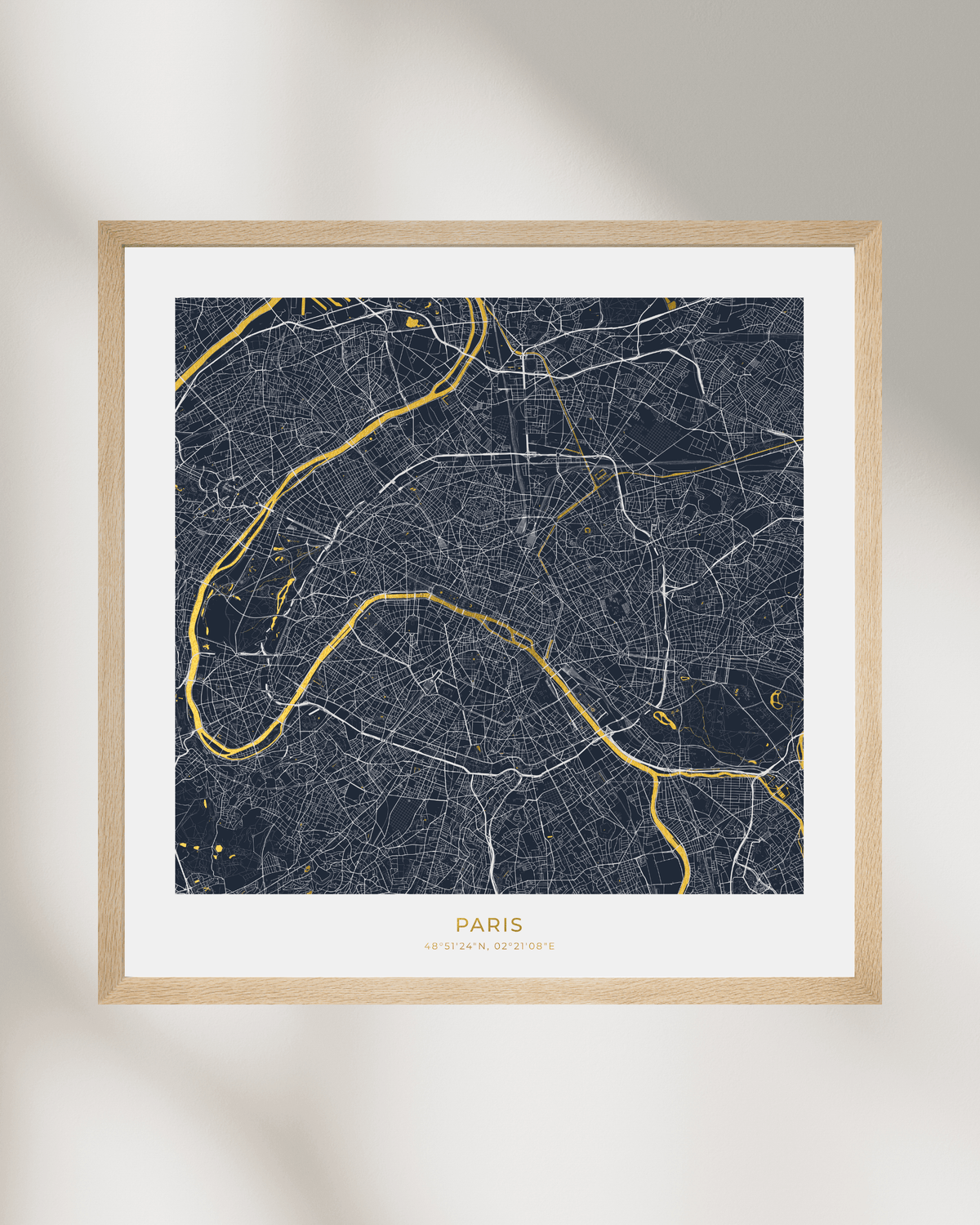 A framed Paris city map art print with a light wood frame, white mat, and navy background with gold street lines and text, shown against a light wall with soft shadows.