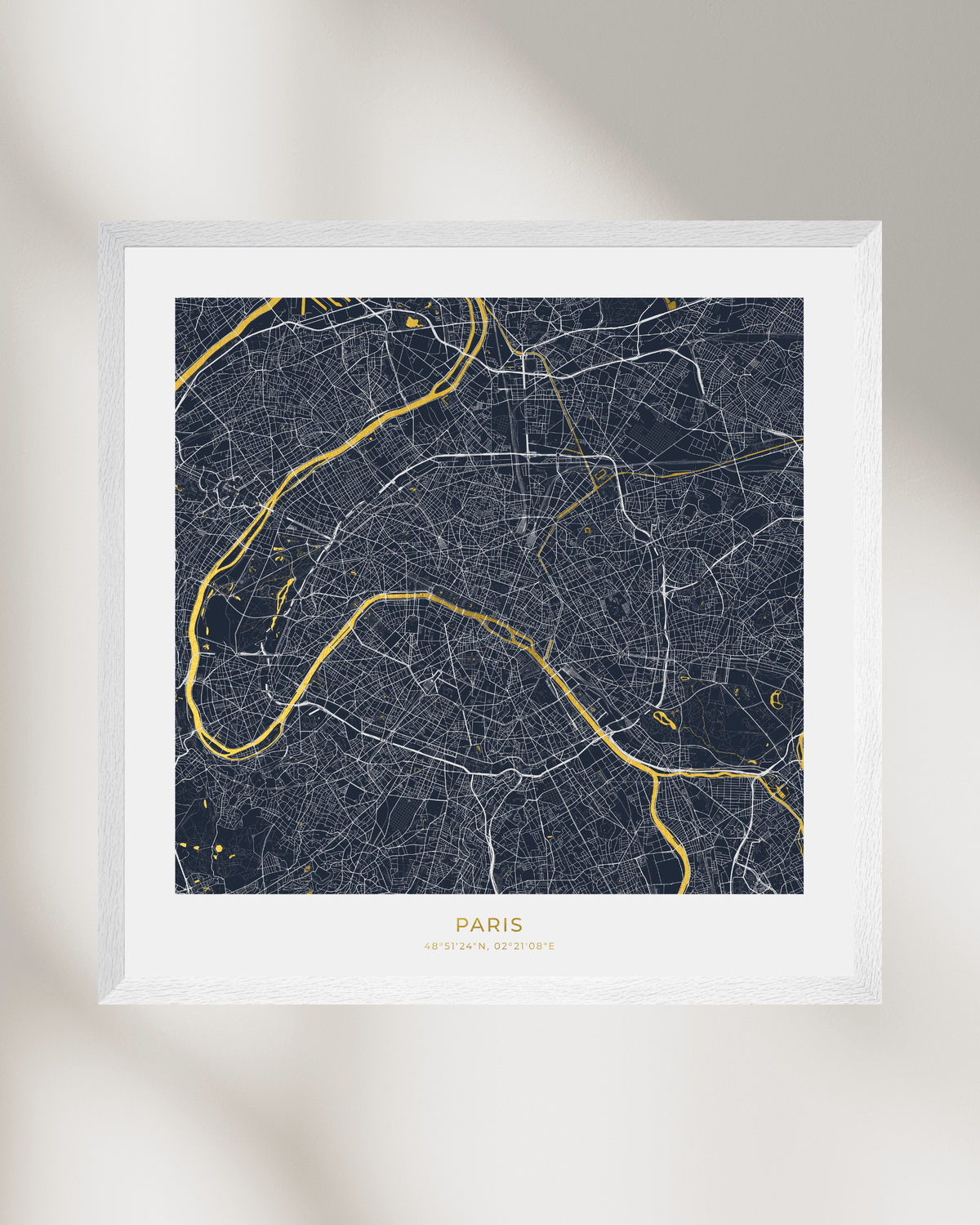A framed Paris city map art print with a dark navy background, white street lines, and gold river and text accents, shown against a light neutral wall with soft shadows.