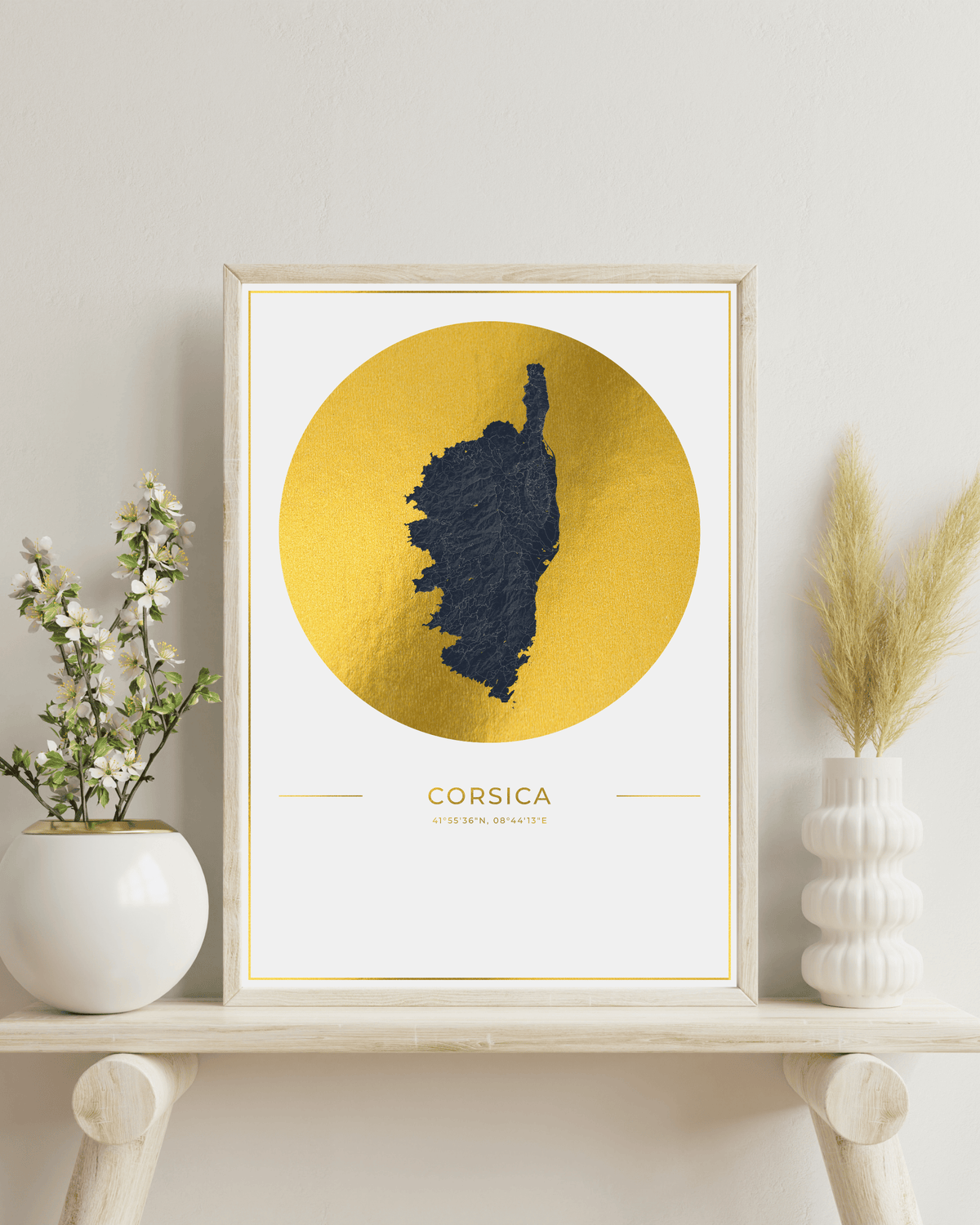 A framed art print featuring a dark blue silhouette map of Corsica inside a large textured gold circle on a white background, styled on a light wood shelf with decorative vases.