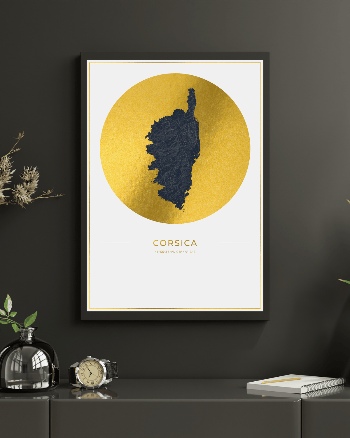 A framed minimalist map print of Corsica hangs on a dark charcoal wall above a console table. The print features a textured dark blue map silhouette centered within a large metallic gold circle, with 'CORSICA'  printed below in gold.
