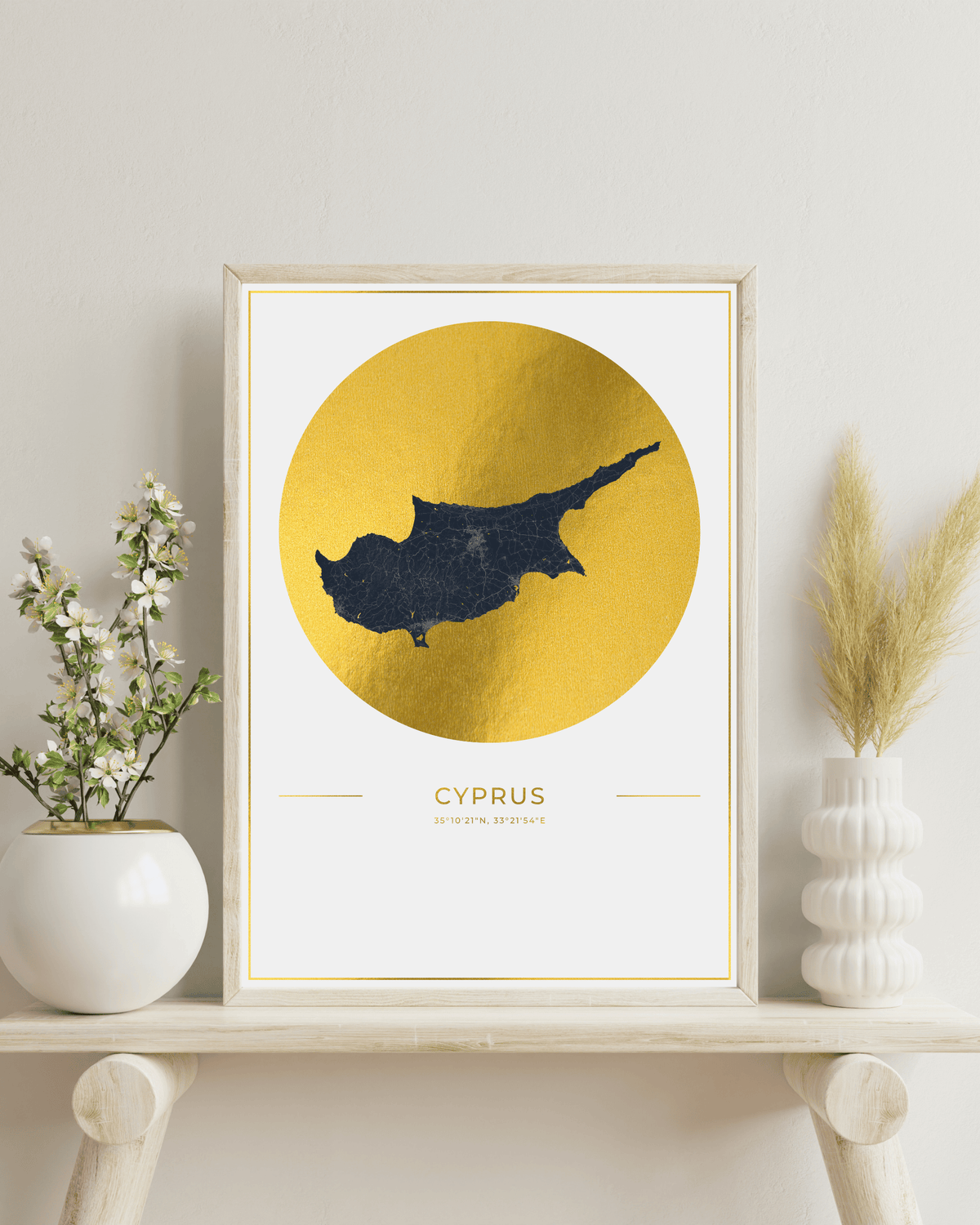 A framed art print featuring a dark navy blue silhouette map of Cyprus inside a large textured gold circle on a white background. The print is styled on a light wood shelf next to a white ceramic vase with small white flowers and a white textured vase with dried pampas grass, against a neutral light gray wall.