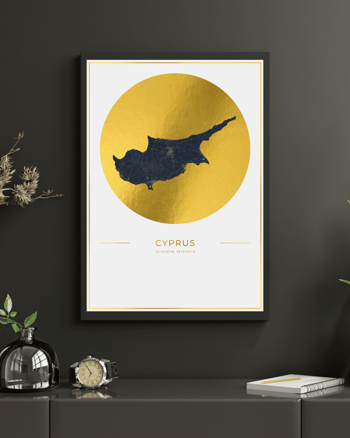 Framed wall art featuring a dark blue silhouette map of Cyprus inside a large textured gold circle on a white background, with the word 'CYPRUS' and small decorative lines below. The frame hangs on a dark charcoal wall above a console table with a glass bottle, clock, and notebook.