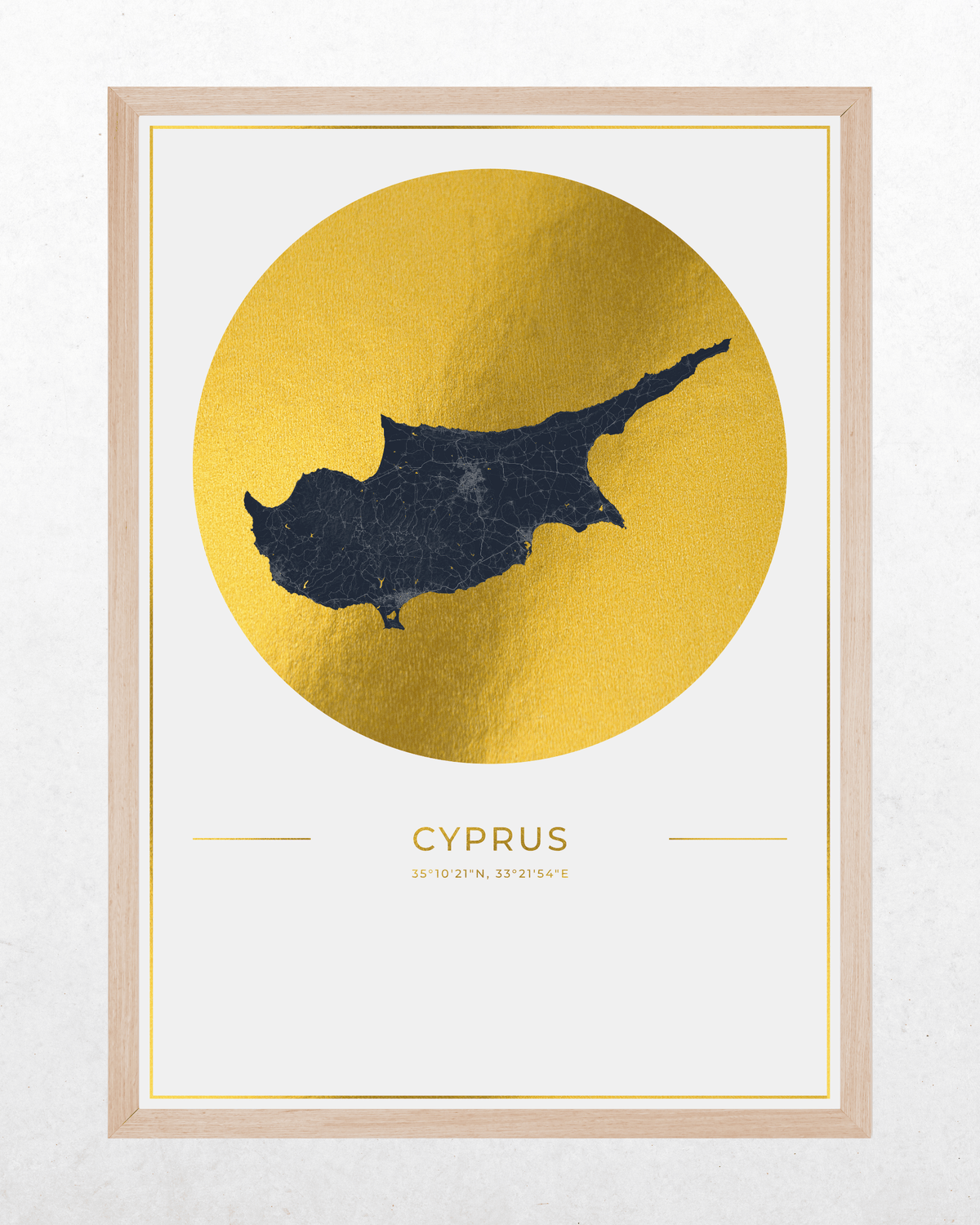 Framed wall art print featuring a dark navy map of Cyprus centered inside a large textured gold circle, with the word "CYPRUS" and small location text below. The print is set in a light natural wood frame against a light neutral wall.