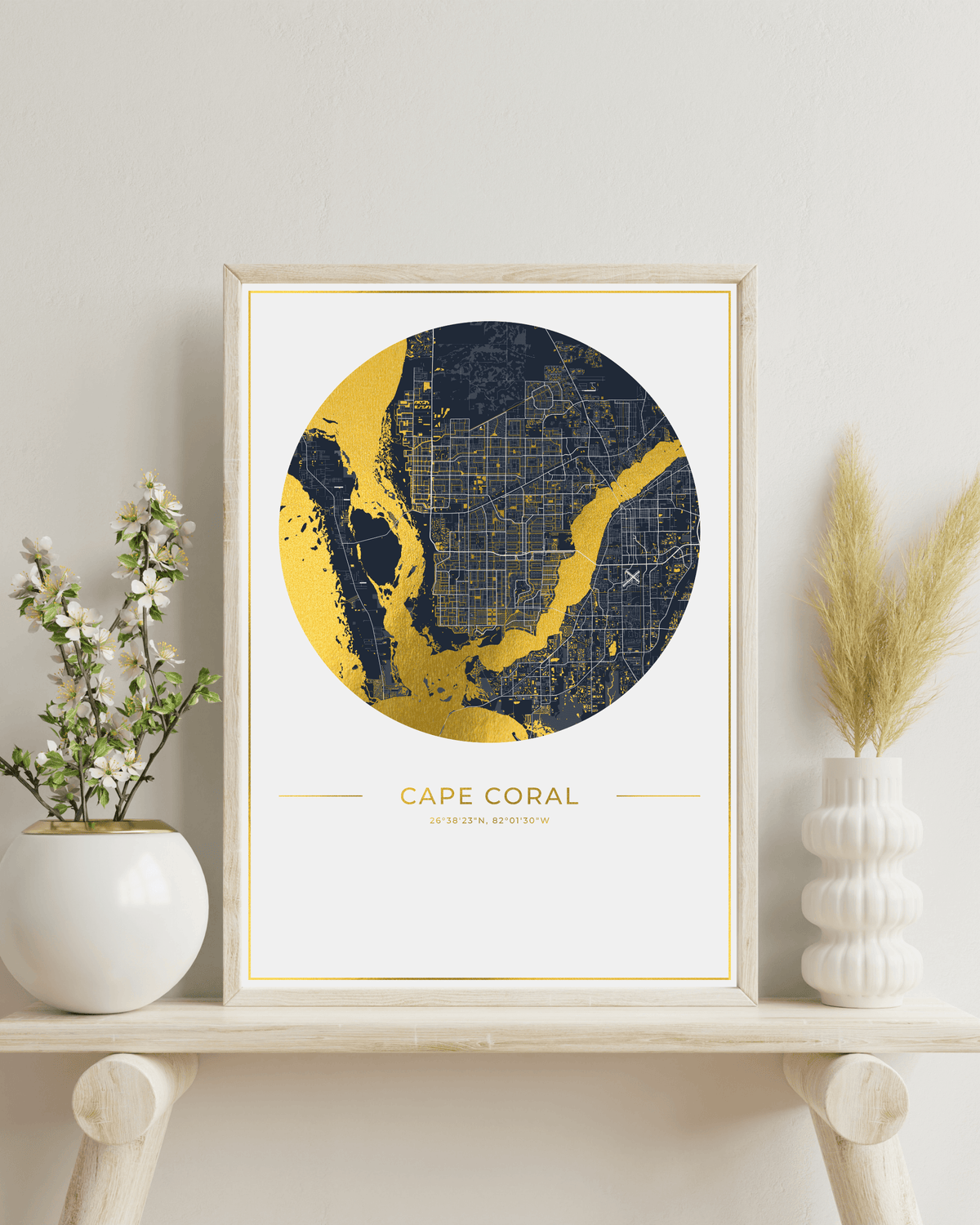 A framed map print of Cape Coral, Florida, featuring a navy blue and gold circular map design with a light wood frame, styled on a wooden shelf with white ceramic vases and dried grass.
