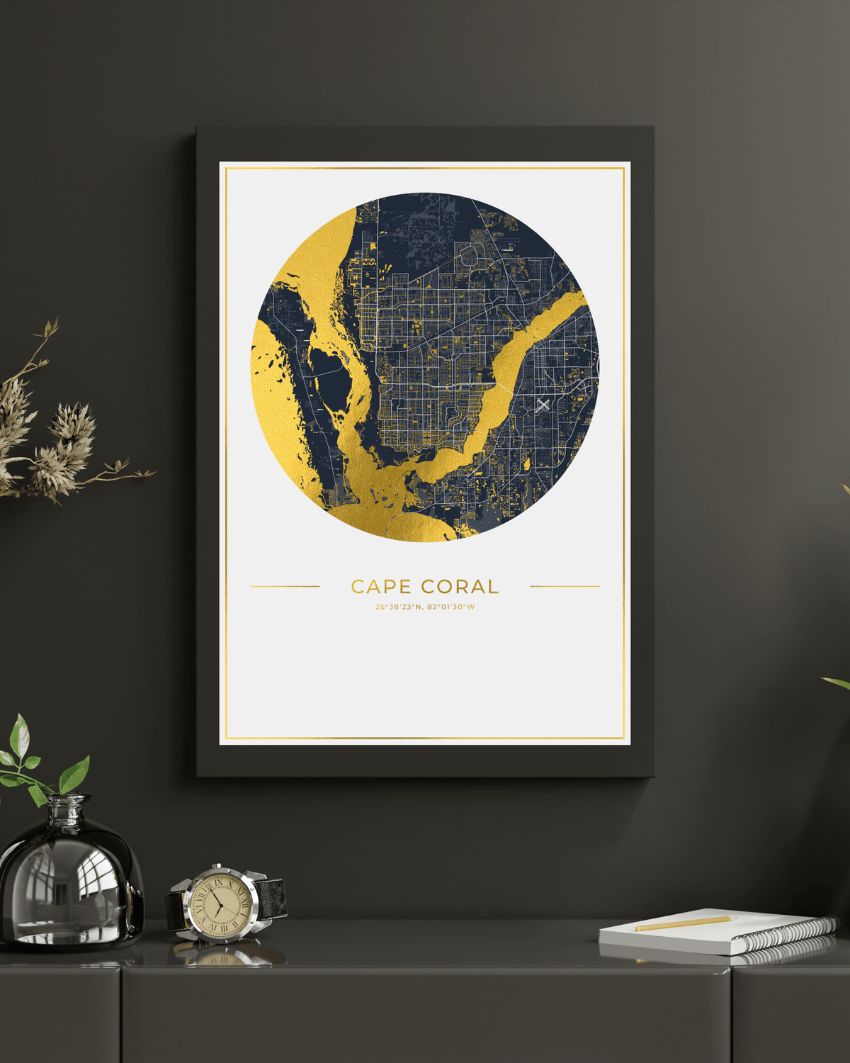 A framed art print of a Cape Coral map, featuring a circular graphic in navy blue and gold on a white background, styled on a dark gray wall above a console table with a clock, glass bottle, and notebook.